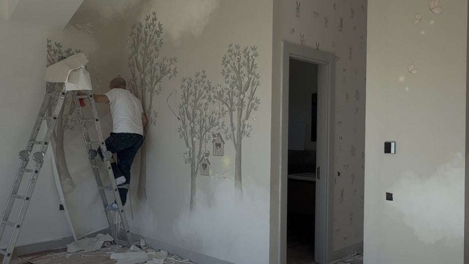 Eski duvar kağıdı sökülmüş çocuk odasında usta, ölçüye özel üretilmiş ağaç desenli yeni duvar kağıdını uyguluyor; zeminde sökülen kağıt şeritleri