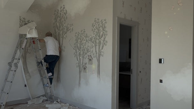 Eski duvar kağıdı sökülmüş çocuk odasında usta, ölçüye özel üretilmiş ağaç desenli yeni duvar kağıdını uyguluyor; zeminde sökülen kağıt şeritleri