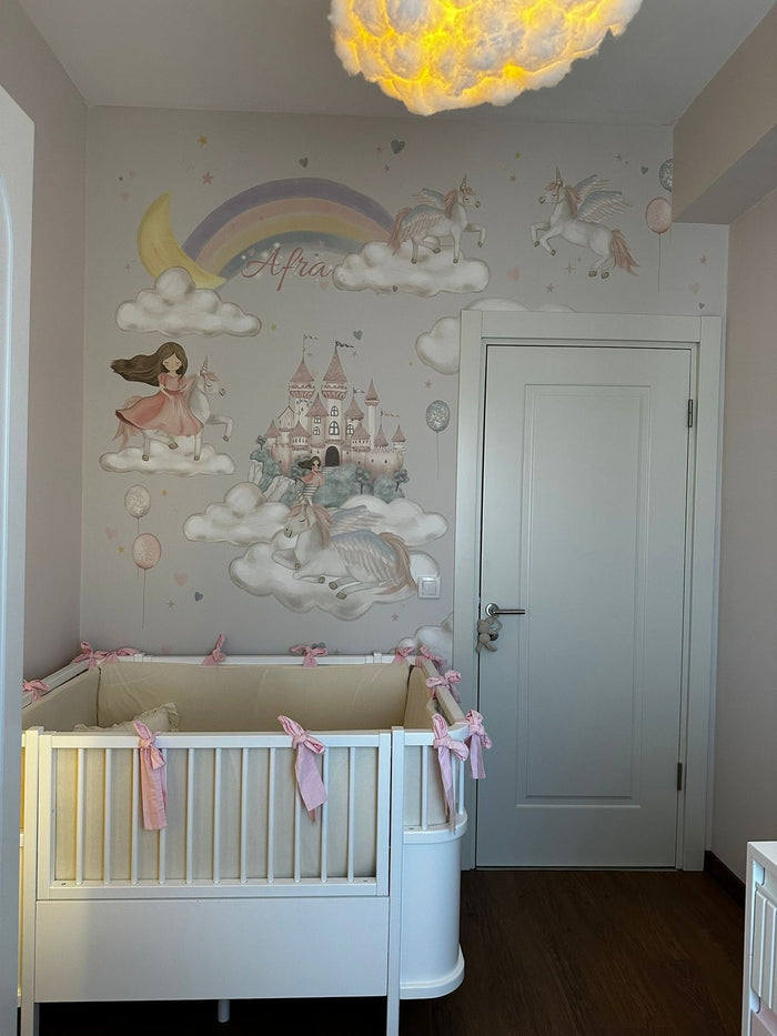 Bebek odasında bulutlar üstünde kanatlı unicornlar, ay ve gökkuşağı, şato ve unicorna binen kız; önde beyaz karyola, yanda kapı